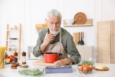 Photo of Elderly man using tablet while cooking at white marble table in kitchen