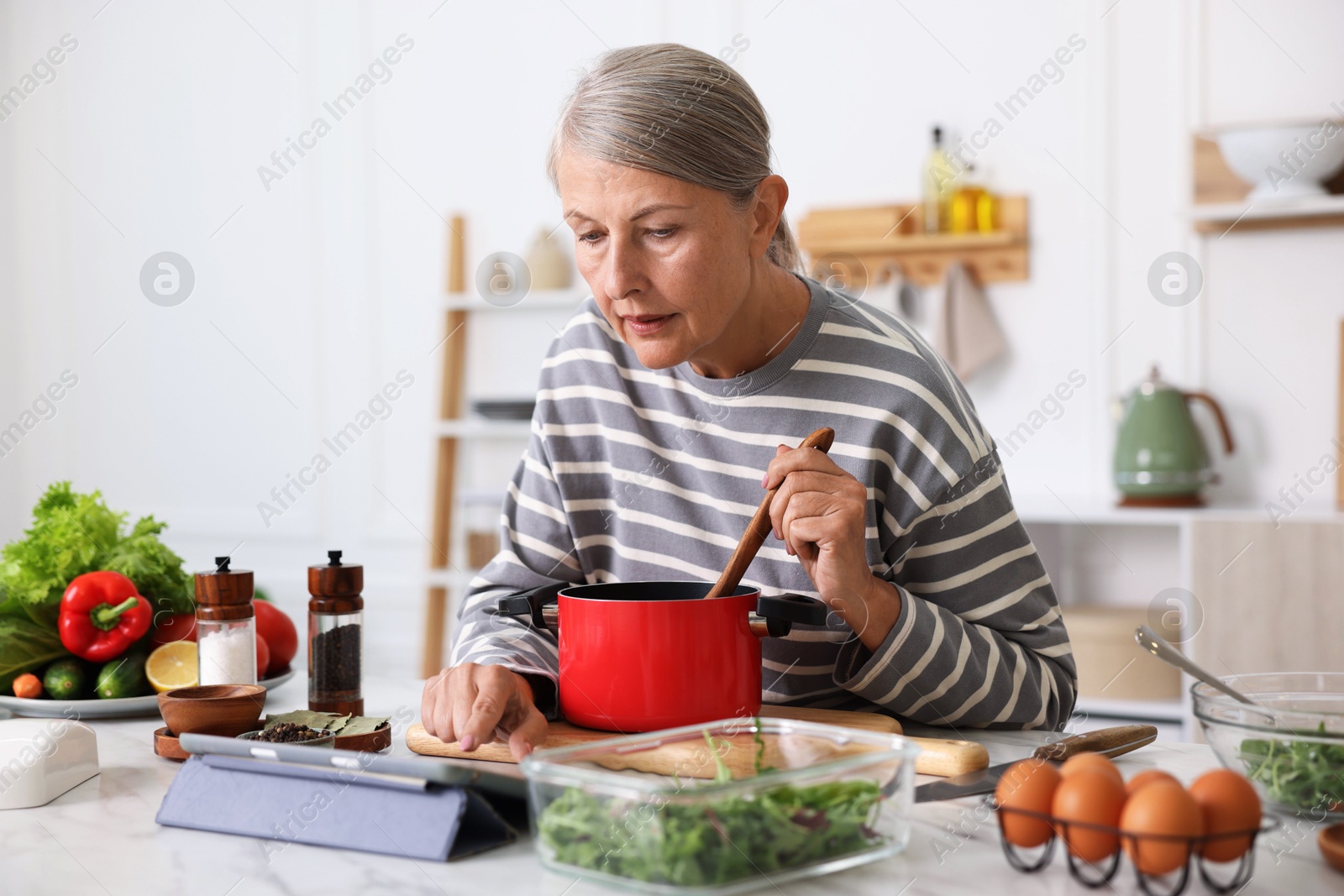 Senior woman using tablet while cooking at white marble table in kitchen Photo of Senior woman using tablet while cooking at white marble table in kitchen