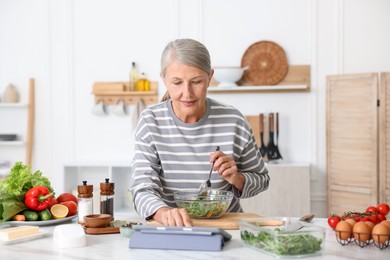 Photo of Senior woman using tablet while cooking at white marble table in kitchen