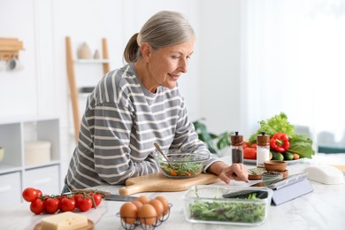 Photo of Senior woman using tablet while cooking at white marble table in kitchen