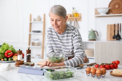 Photo of Senior woman using tablet while cooking at white marble table in kitchen