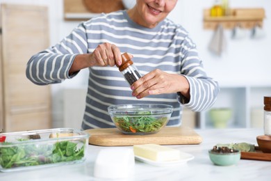 Photo of Cooking process. Senior woman adding spices to salad at white marble table in kitchen, closeup