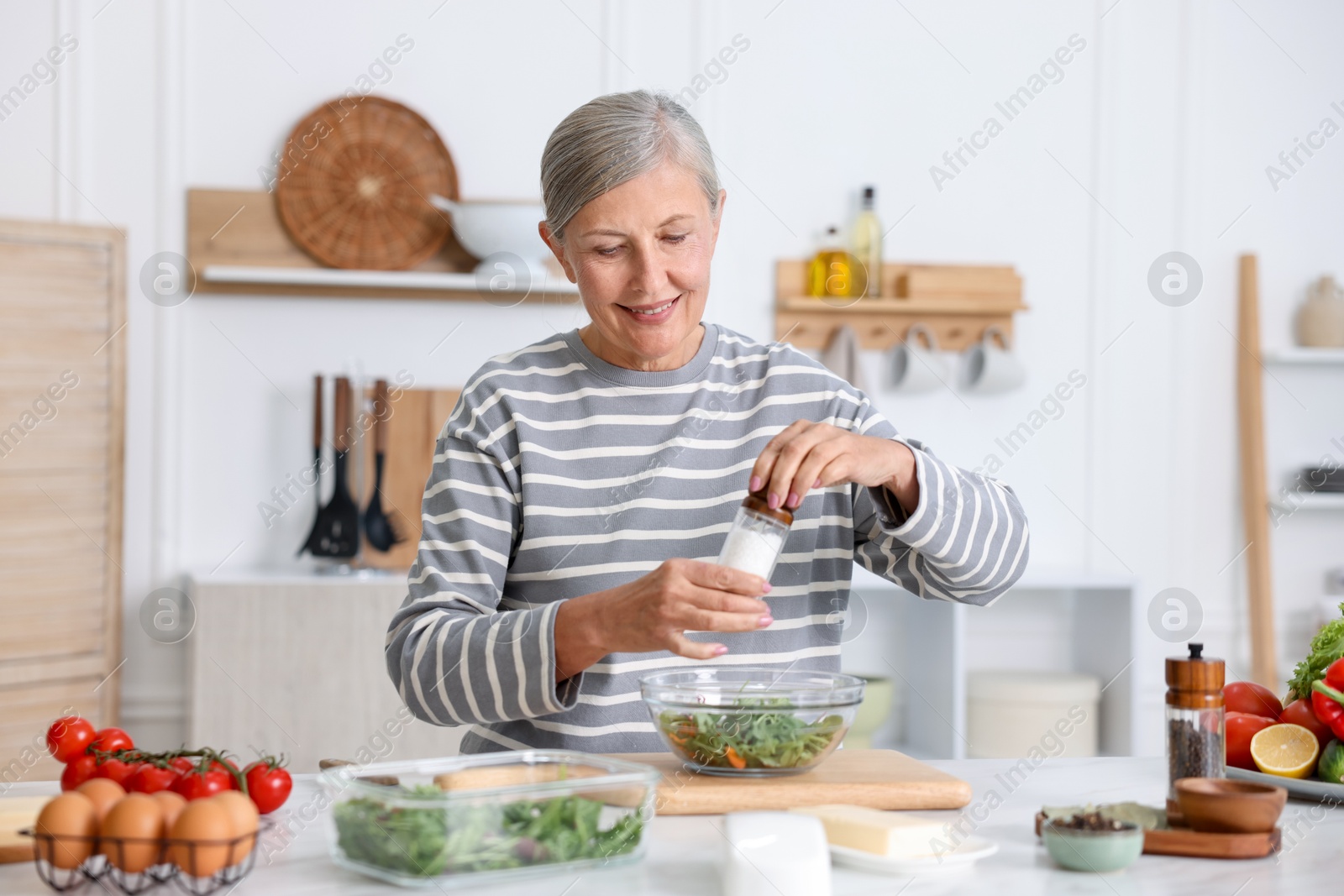 Cooking process. Senior woman adding spices to salad at white marble table in kitchen Photo of Cooking process. Senior woman adding spices to salad at white marble table in kitchen