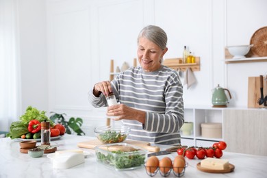 Photo of Cooking process. Senior woman adding spices to salad at white marble table in kitchen
