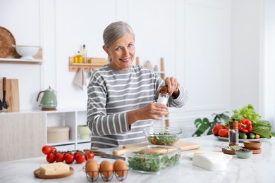 Photo of Cooking process. Senior woman adding spices to salad at white marble table in kitchen