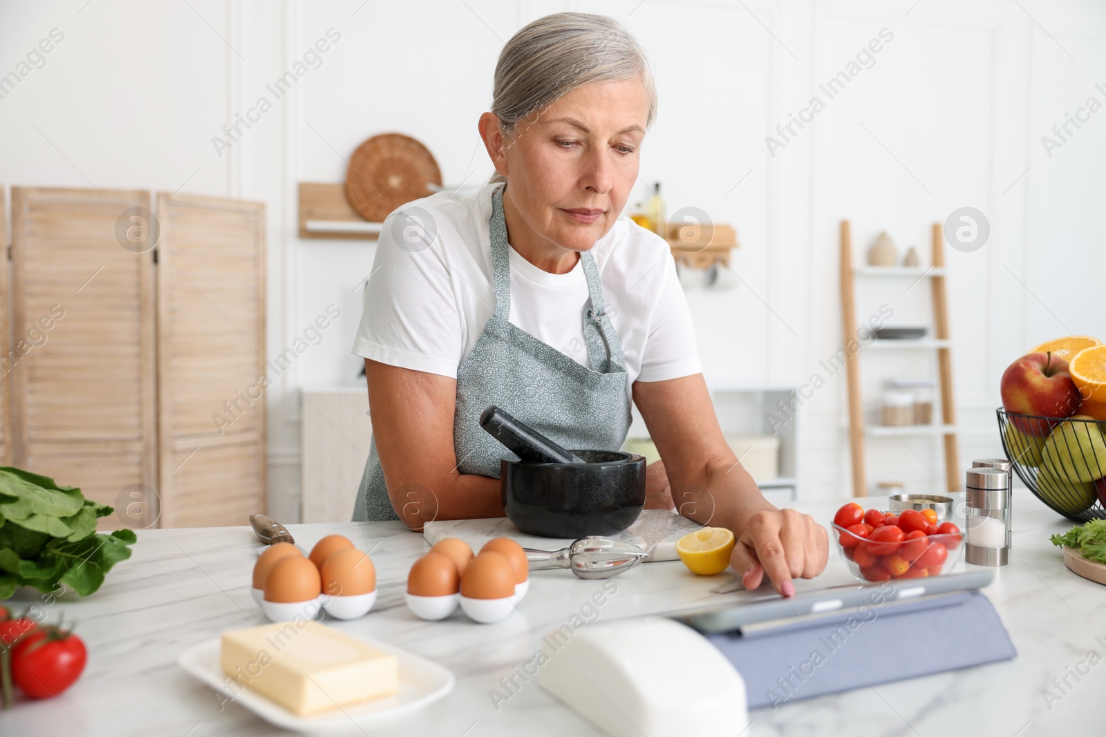 Senior woman using tablet while cooking at white marble table in kitchen Photo of Senior woman using tablet while cooking at white marble table in kitchen