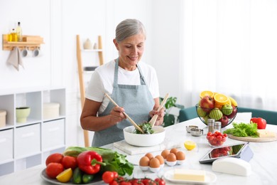 Photo of Senior woman cooking salad at white marble table in kitchen
