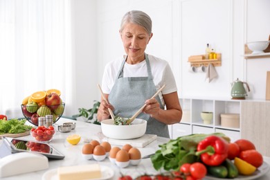 Photo of Senior woman cooking salad at white marble table in kitchen