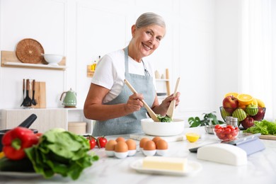 Photo of Senior woman cooking salad at white marble table in kitchen