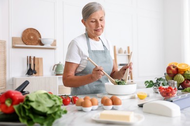 Photo of Senior woman cooking salad at white marble table in kitchen