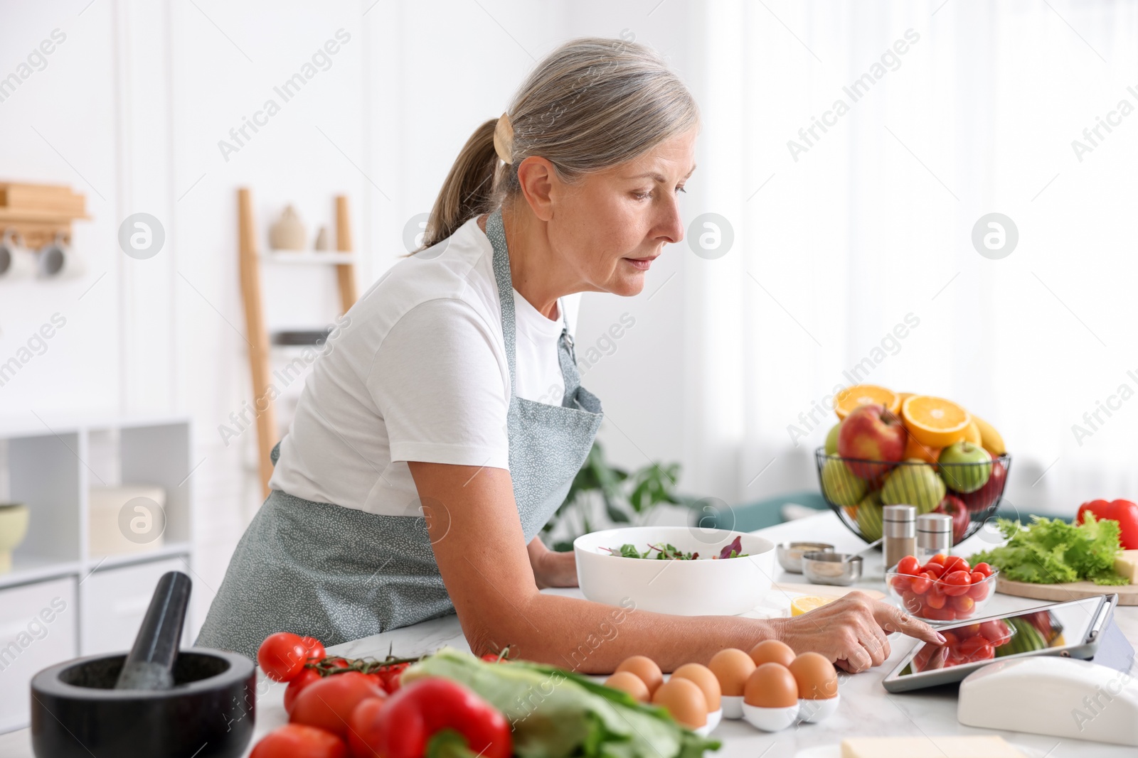 Senior woman using tablet while cooking salad at white marble table in kitchen Photo of Senior woman using tablet while cooking salad at white marble table in kitchen