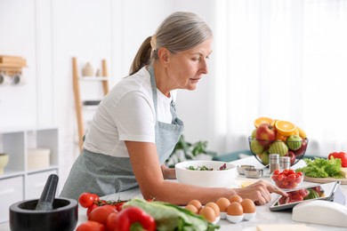 Photo of Senior woman using tablet while cooking salad at white marble table in kitchen