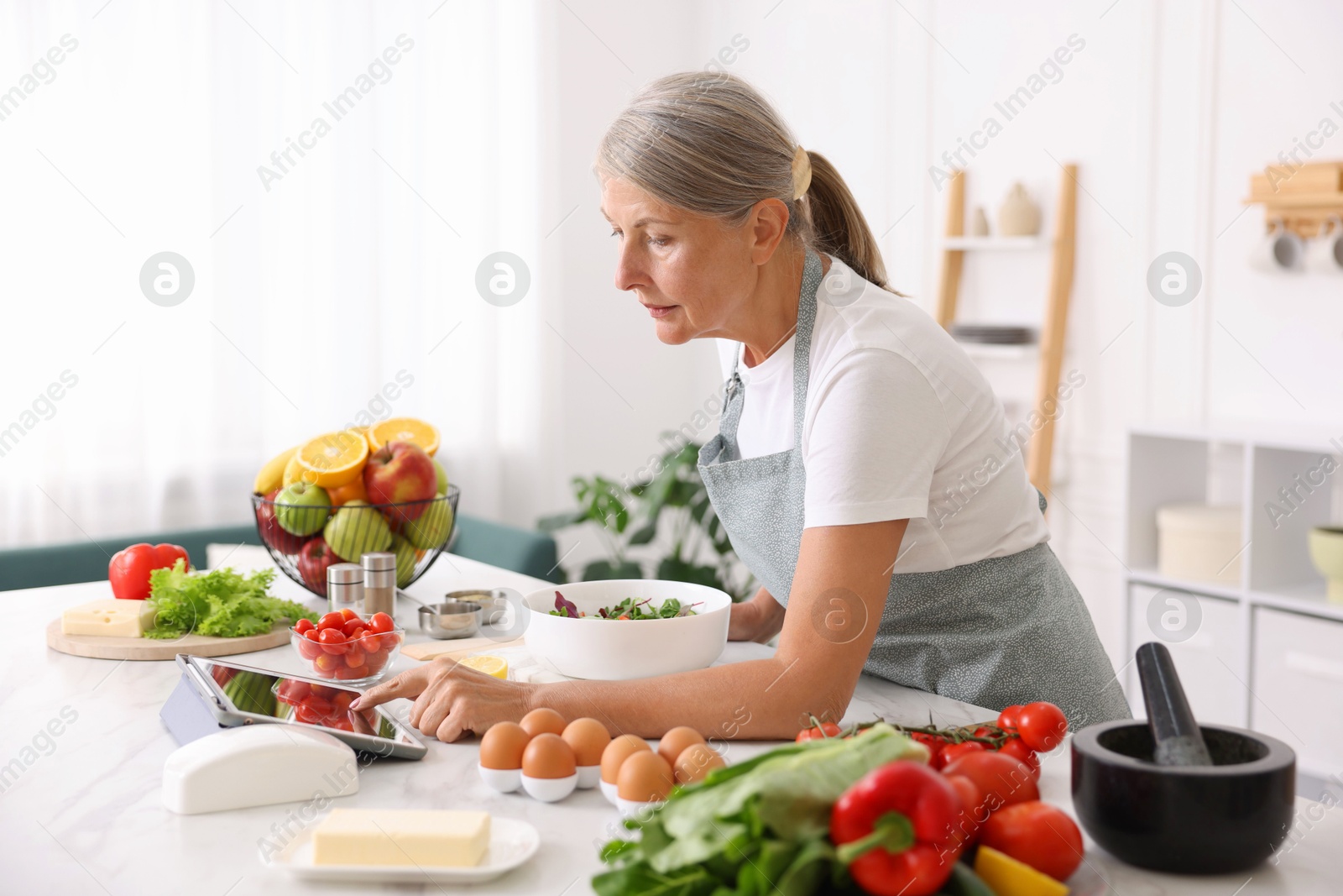Senior woman using tablet while cooking salad at white marble table in kitchen Photo of Senior woman using tablet while cooking salad at white marble table in kitchen