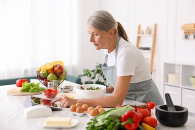 Photo of Senior woman using tablet while cooking salad at white marble table in kitchen