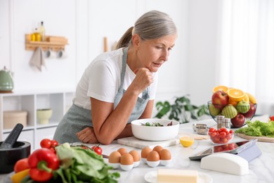 Photo of Senior woman using tablet while cooking salad at white marble table in kitchen