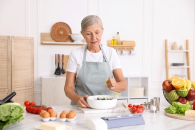 Photo of Senior woman cooking salad at white marble table in kitchen