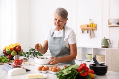 Photo of Senior woman cooking salad at white marble table in kitchen