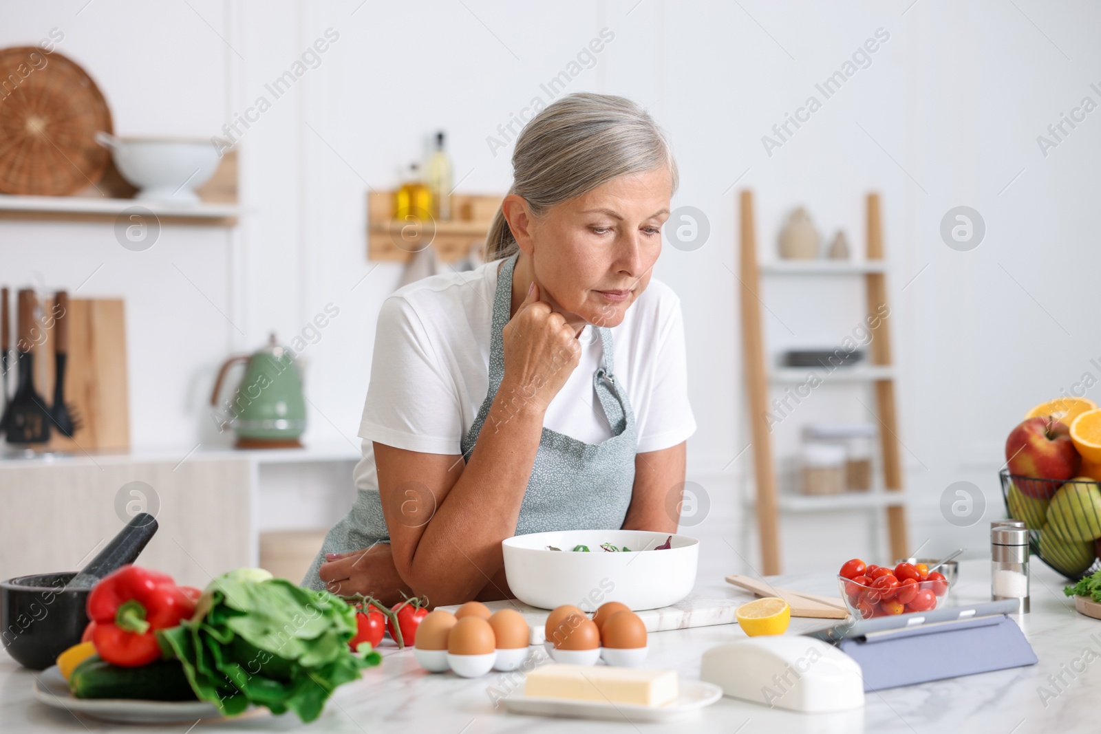 Senior woman using tablet while cooking salad at white marble table in kitchen Photo of Senior woman using tablet while cooking salad at white marble table in kitchen