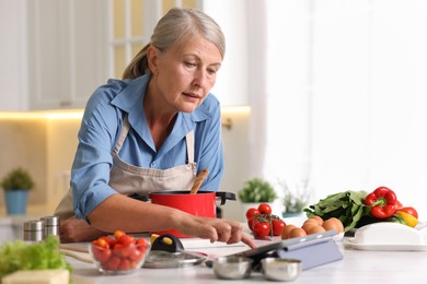 Photo of Senior woman using tablet while cooking at white marble table in kitchen