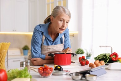 Photo of Senior woman using tablet while cooking at white marble table in kitchen