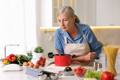 Photo of Senior woman using tablet while cooking at white marble table in kitchen