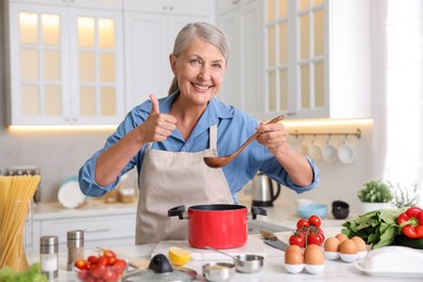 Photo of Senior woman showing thumbs up while cooking at white marble table in kitchen