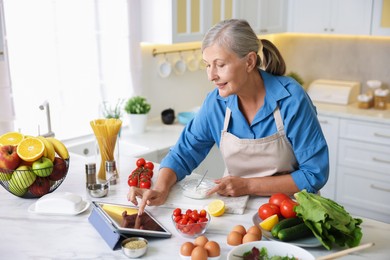 Photo of Senior woman using tablet while cooking at white marble table in kitchen