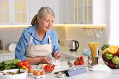 Photo of Senior woman using tablet while cooking at white marble table in kitchen