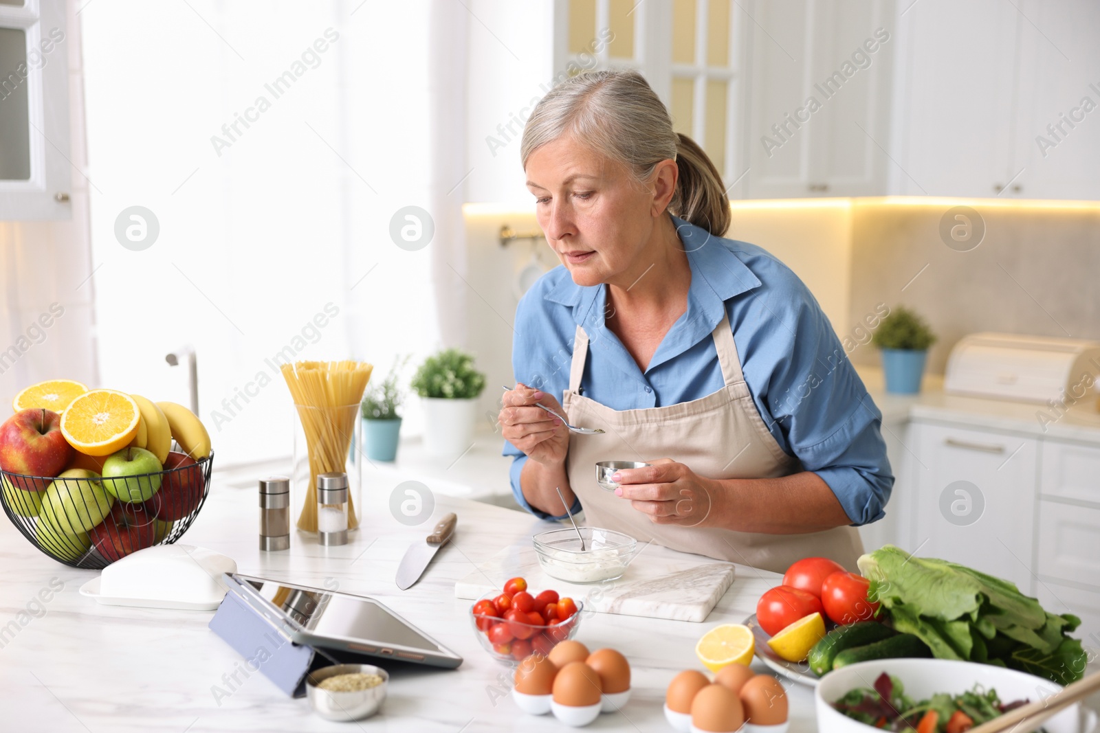 Senior woman using tablet while cooking at white marble table in kitchen Photo of Senior woman using tablet while cooking at white marble table in kitchen