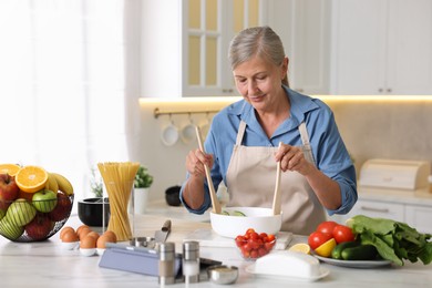 Photo of Senior woman cooking salad at white marble table in kitchen