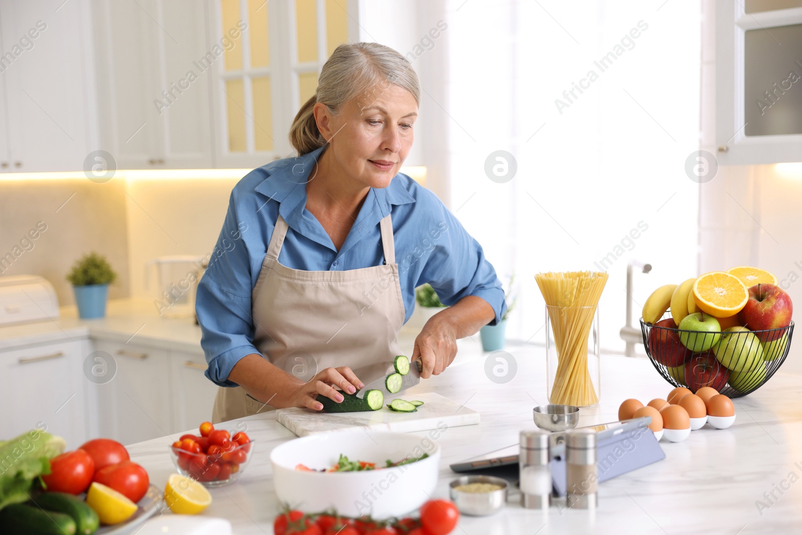 Senior woman using tablet while cooking at white marble table in kitchen Photo of Senior woman using tablet while cooking at white marble table in kitchen