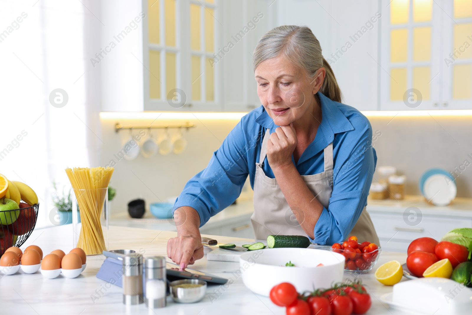 Senior woman using tablet while cooking at white marble table in kitchen Photo of Senior woman using tablet while cooking at white marble table in kitchen