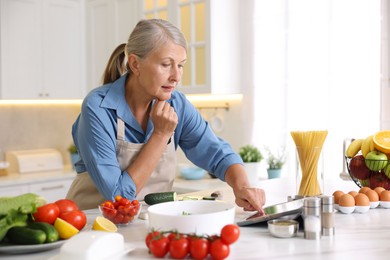 Photo of Senior woman using tablet while cooking at white marble table in kitchen