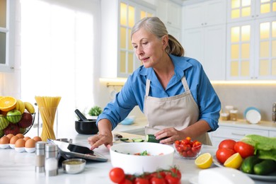 Photo of Senior woman using tablet while cooking at white marble table in kitchen