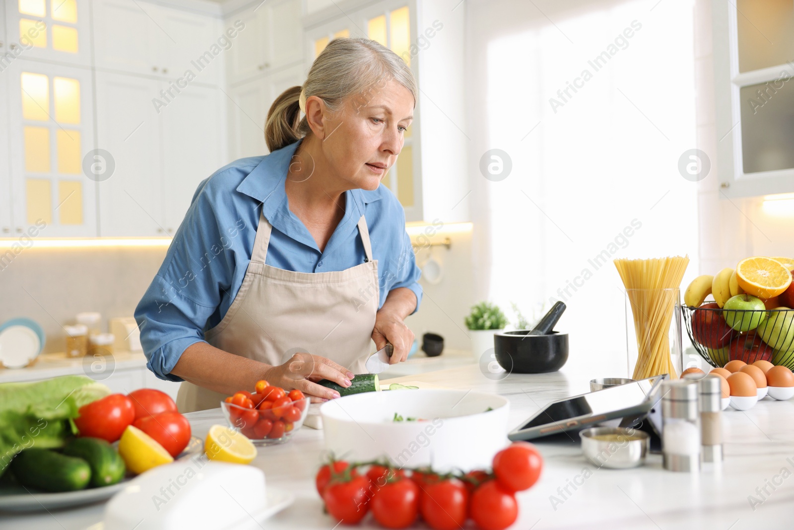 Senior woman using tablet while cooking at white marble table in kitchen Photo of Senior woman using tablet while cooking at white marble table in kitchen