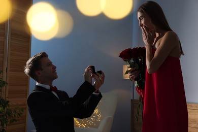 Photo of Smiling man with engagement ring making marriage proposal to his excited girlfriend in dark room. Bokeh effect