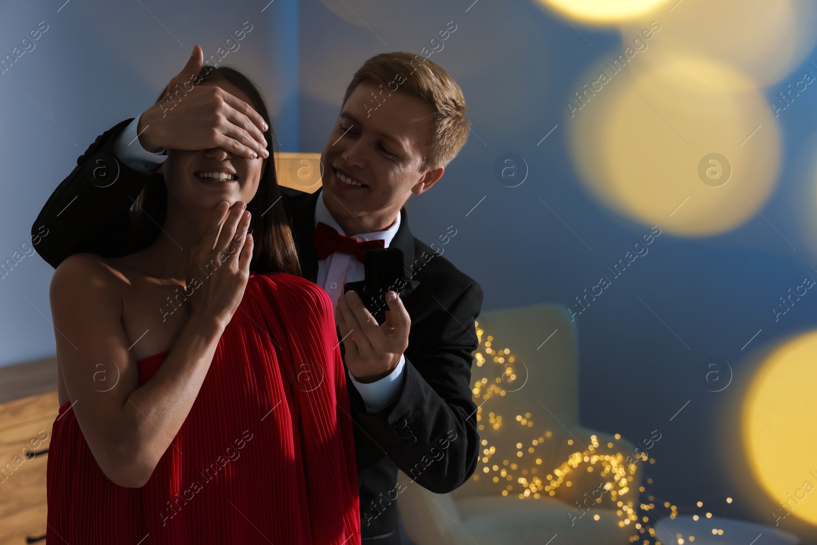 Making marriage proposal. Smiling man with engagement ring surprising his girlfriend in dark room, space for text. Bokeh effect Photo of Making marriage proposal. Smiling man with engagement ring surprising his girlfriend in dark room, space for text. Bokeh effect