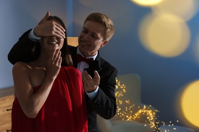 Photo of Making marriage proposal. Smiling man with engagement ring surprising his girlfriend in dark room, space for text. Bokeh effect