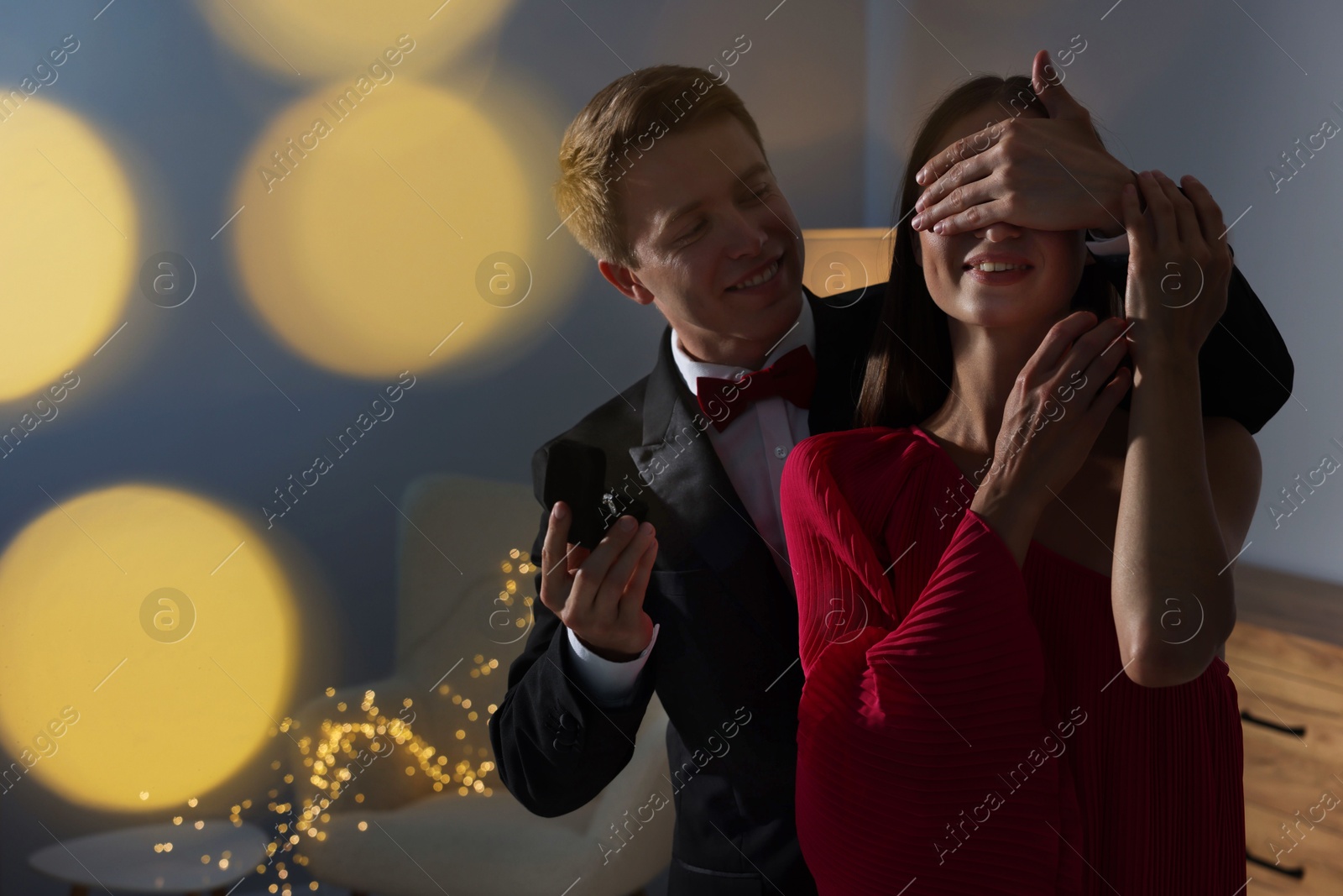 Making marriage proposal. Smiling man with engagement ring surprising his girlfriend in dark room, space for text. Bokeh effect Photo of Making marriage proposal. Smiling man with engagement ring surprising his girlfriend in dark room, space for text. Bokeh effect