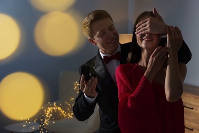 Photo of Making marriage proposal. Smiling man with engagement ring surprising his girlfriend in dark room, space for text. Bokeh effect