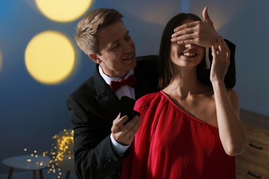 Photo of Making marriage proposal. Smiling man with engagement ring surprising his girlfriend in dark room. Bokeh effect