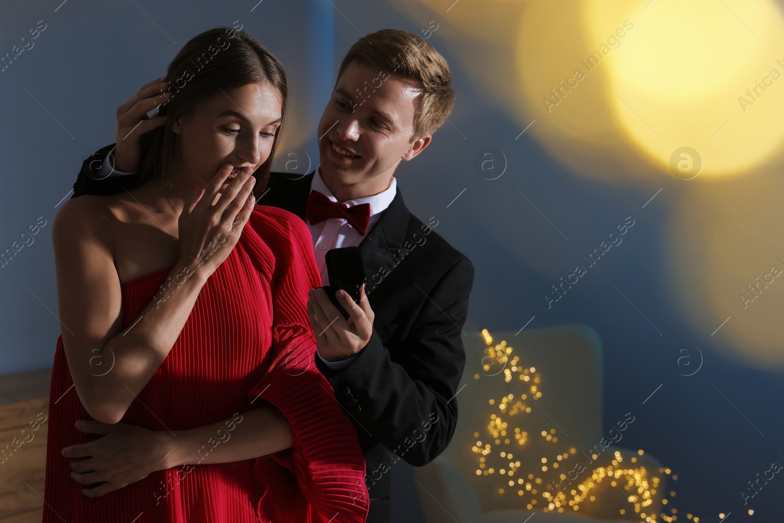 Making marriage proposal. Smiling man with engagement ring surprising his girlfriend in dark room, space for text. Bokeh effect Photo of Making marriage proposal. Smiling man with engagement ring surprising his girlfriend in dark room, space for text. Bokeh effect