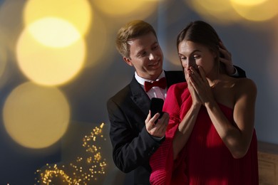 Photo of Making marriage proposal. Smiling man with engagement ring surprising his girlfriend in dark room, space for text. Bokeh effect