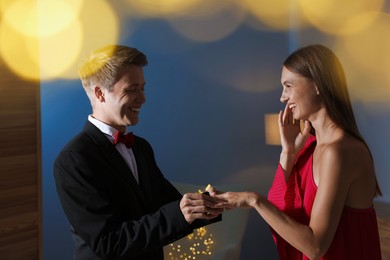 Photo of Making marriage proposal. Smiling man putting engagement ring onto his girlfriend's finger in dark room. Bokeh effect