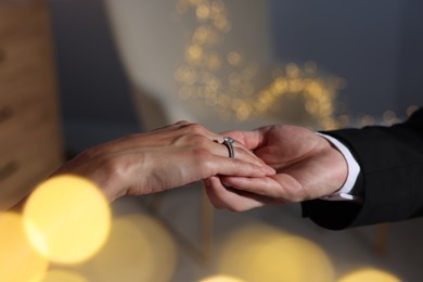 Photo of Making marriage proposal. Woman with engagement ring and her fiance holding hands at home, closeup. Bokeh effect