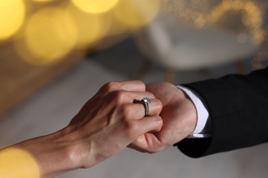 Photo of Making marriage proposal. Woman with engagement ring and her fiance holding hands at home, closeup. Bokeh effect