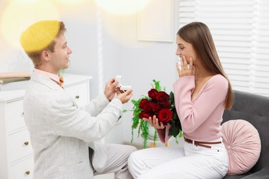 Photo of Man with engagement ring and bouquet of roses making marriage proposal to his emotional girlfriend at home. Bokeh effect