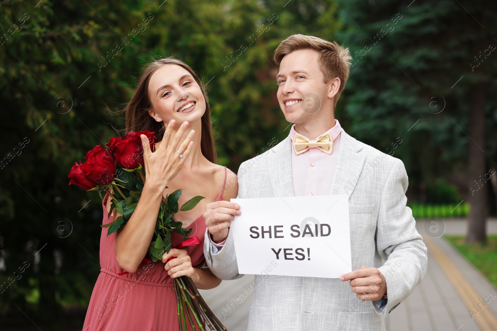 Smiling man holding sheet of paper with text She Said Yes and his happy beloved with bouquet of roses showing ring on engagement day outdoors Photo of Smiling man holding sheet of paper with text She Said Yes and his happy beloved with bouquet of roses showing ring on engagement day outdoors