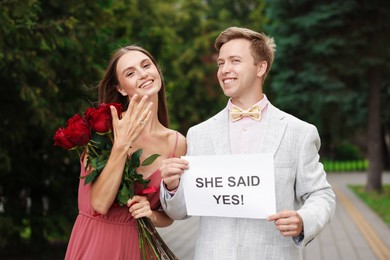 Photo of Smiling man holding sheet of paper with text She Said Yes and his happy beloved with bouquet of roses showing ring on engagement day outdoors
