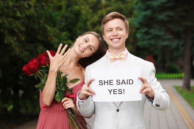 Photo of Smiling man holding sheet of paper with text She Said Yes and his happy beloved with bouquet of roses showing ring on engagement day outdoors